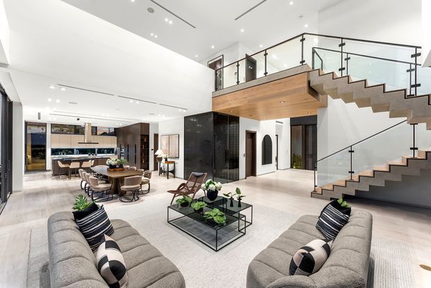 A bright, expansive open-plan contemporary interior combining a living room, dining area, and kitchen beneath a high ceiling. Two large grey sofas face a glass coffee table, while a dining table and kitchen sit beyond. A dramatic timber-and-glass staircase rises to a mezzanine level, with black-framed glazing and sleek dark built-in cabinetry creating strong contrast against the white walls and pale flooring.
