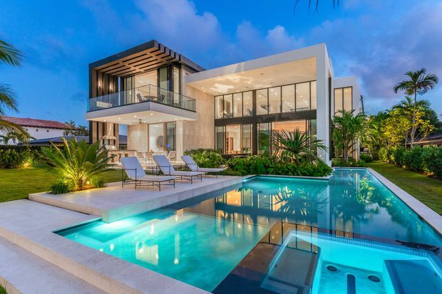 Exterior dusk view of a large contemporary two-storey residence with extensive floor-to-ceiling glazing, projecting upper-level balcony, timber-look pergola screening, landscaped tropical garden, paved pool terrace, and a large illuminated swimming pool in the foreground.