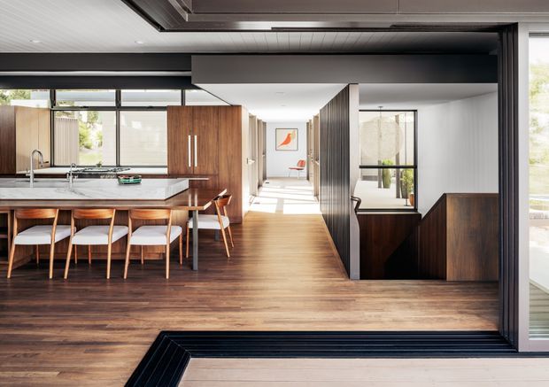 A bright, open-plan contemporary interior centred on a timber kitchen and dining area. The kitchen features a large white stone island with integrated sink and seating for several people, dark timber cabinetry, and extensive glazing. Warm timber flooring continues toward a stair opening framed in dark timber, while a black-framed internal partition and layered ceiling detail add architectural definition.