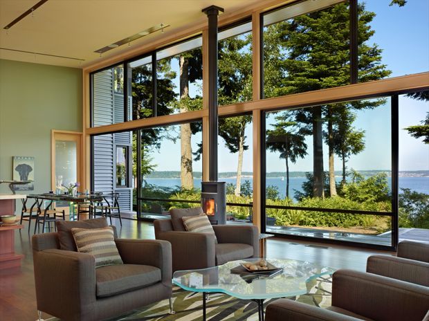 A spacious contemporary living room with floor-to-ceiling timber-framed glazing overlooking a wooded waterfront. The room features multiple upholstered armchairs, a glass coffee table, a dining area, polished timber flooring, and a compact freestanding fireplace. Abundant natural light and expansive views create an open, relaxed atmosphere.