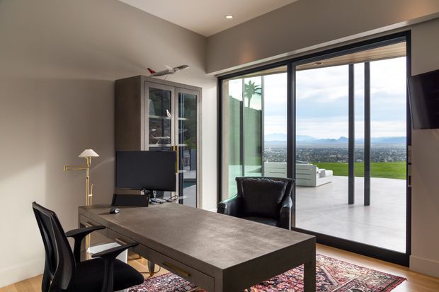 A refined home office with a large dark wood or stone-look desk, ergonomic black chairs, a glass-front display cabinet, floor lamp, recessed ceiling lighting, and a wall-mounted screen. Full-height black-framed glazing opens visually onto an outdoor terrace and expansive distant landscape, bringing in abundant natural light.