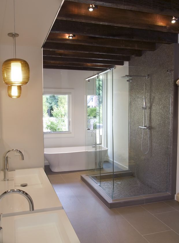 A spacious contemporary bathroom with a freestanding white bathtub, double vanity, and large frameless glass-enclosed walk-in shower. The shower and adjacent wall are finished in small dark mosaic tiles, while the floor uses large taupe-grey tiles. A dark exposed timber-beam ceiling contrasts with pale walls and white sanitaryware. Warm pendant and recessed lighting create a refined, softly atmospheric interior.