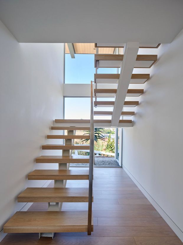 A bright, minimalist stairway with floating timber treads, a slim central timber handrail, and white structural supports. Large floor-to-ceiling glazing and a roof opening bring in abundant daylight, with views to planted outdoor space beyond. The stair connects to an upper level beneath a light timber-lined ceiling.