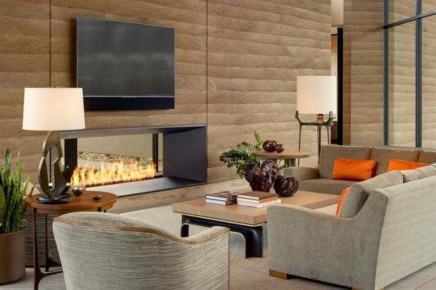 A refined contemporary living room featuring a large sectional sofa and armchairs arranged around a low coffee table, with a built-in linear fireplace beneath a wall-mounted television. The room has warm textured wood wall panelling, dark built-in fireplace cabinetry, floor-to-ceiling glazing, table and floor lamps, and orange accent cushions. The overall atmosphere is warm, luxurious, and inviting.
