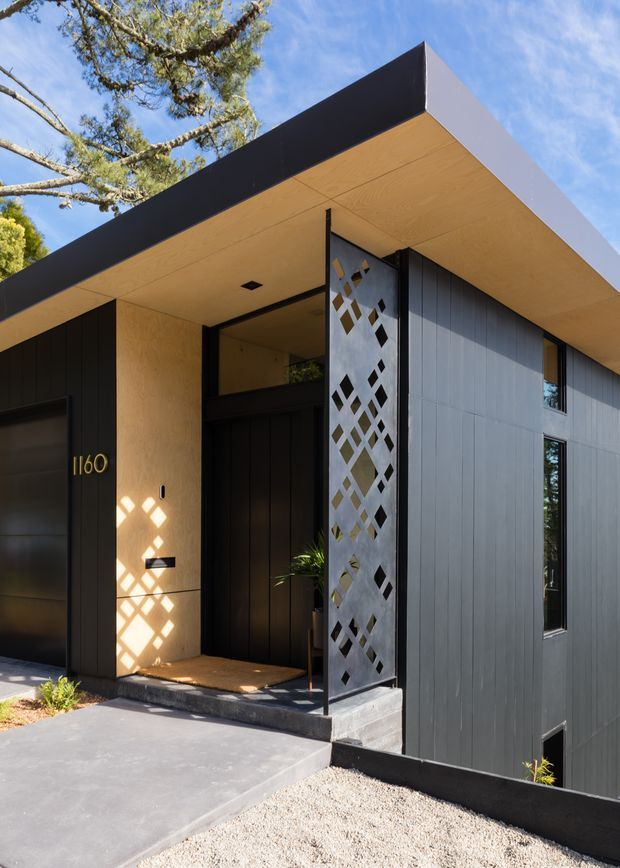 Exterior view of a contemporary single-storey residential entry with a strong flat-roofed form, deep timber-lined soffit, dark vertical cladding, and a perforated metal privacy screen beside the recessed front door. A concrete driveway and gravel landscape lead to the entry, while narrow vertical windows punctuate the dark facade. The composition is angular and minimalist, with bold black and warm timber tones.