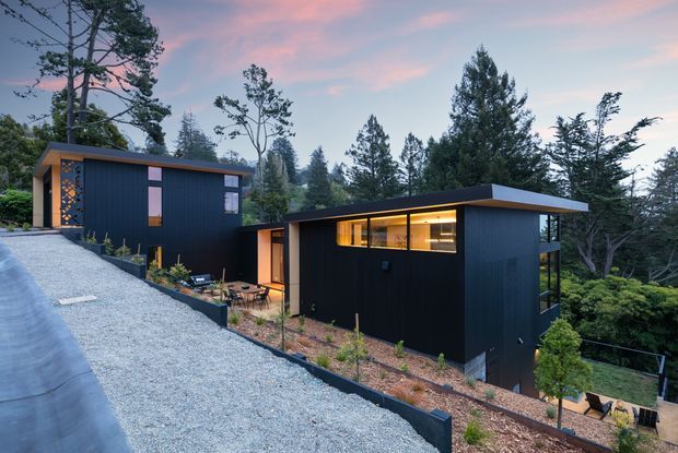Exterior view of a contemporary dark-clad multi-level home composed of two flat-roofed rectangular volumes on a sloping, heavily wooded site. Large windows emit warm interior light, while a gravel driveway and landscaped terraces lead around the building. The dusk sky and surrounding trees create a secluded, modern forest setting.