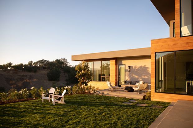 A modern multi-level house viewed from the landscaped garden, featuring warm timber-clad walls, expansive floor-to-ceiling glazing, strong horizontal roof overhangs, and a paved terrace with outdoor lounge seating. The home sits in a quiet landscaped setting with trees and a broad lawn in the foreground, illuminated by warm late-afternoon light.