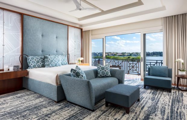 A bright, spacious waterfront bedroom or hotel-style suite with a blue upholstered bed, matching seating area, patterned carpet, and decorative geometric wall panels. Floor-to-ceiling glazing opens to a balcony overlooking a body of water and distant built-up shoreline. The room includes a recessed tray ceiling with a ceiling fan, neutral curtains, and warm timber detailing.