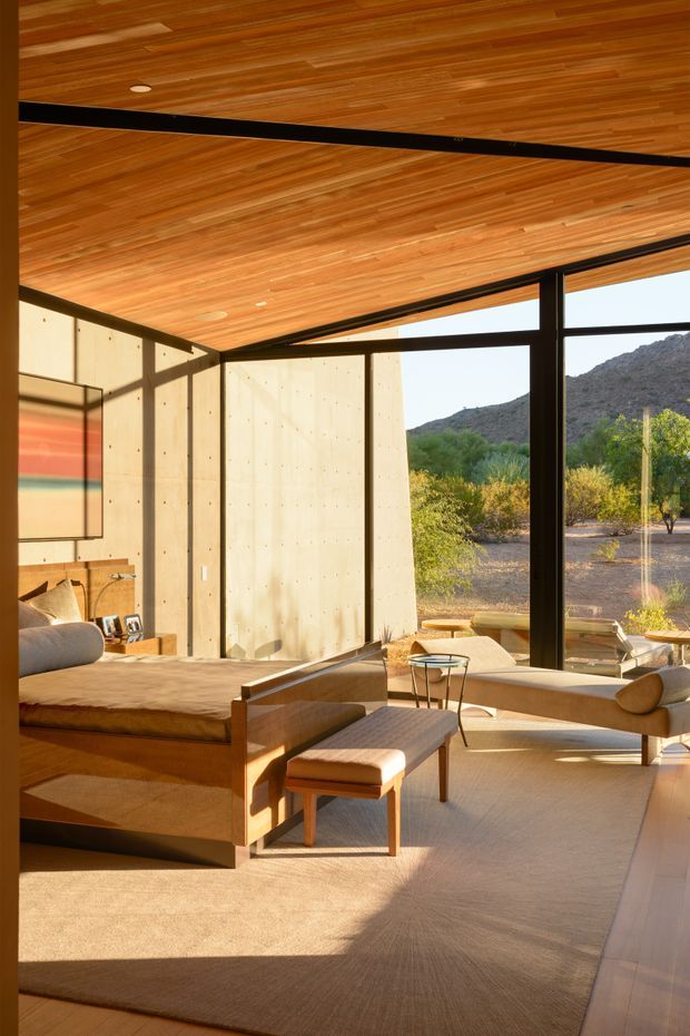 A warm, sunlit mid-century-inspired living room with expansive black-framed glazing overlooking a dry, mountainous landscape. The room features a richly timber-lined ceiling, pale concrete or plaster wall panels, built-in timber cabinetry, a low wooden platform bed or daybed, upholstered lounge seating, a leather or fabric ottoman, and a large neutral rug. Strong horizontal lines, exposed black structural framing, and abundant natural light create a refined, serene interior connected to the landscape.