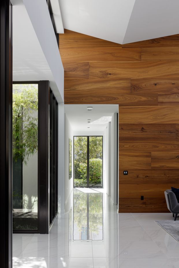 Bright contemporary interior hallway or circulation space with a dramatic full-height walnut timber feature wall, large black-framed glazed openings, white walls and ceiling, and highly reflective pale stone or porcelain flooring. A partial living area with an armchair and rug is visible at right, while greenery is visible through the glazing.
