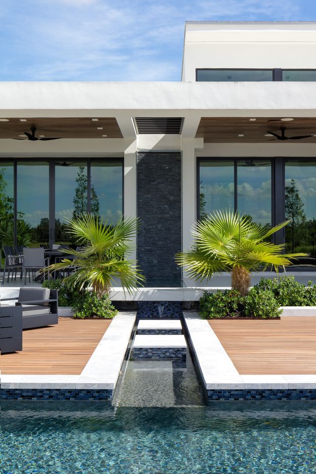 A bright contemporary courtyard pool area viewed toward a modern multi-level residence. The foreground features a swimming pool with a narrow raised water channel and tiled spillway aligned to the building entrance. White rendered walls, dark slate-clad central feature wall, extensive dark-framed glazing, timber decking, ceiling fans, and built-in outdoor seating define the space. Palm plantings and low hedges soften the geometric design.