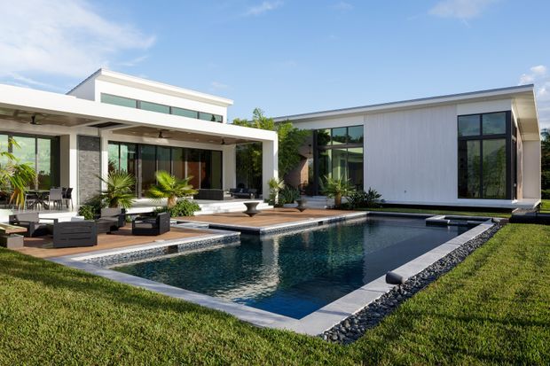 A contemporary multi-wing residence viewed from the landscaped rear yard, with a rectangular swimming pool, broad paved terrace, outdoor lounge furniture, tropical planting, extensive dark-framed glazing, and clean white and grey facade surfaces. The house has predominantly flat roof forms and strong indoor-outdoor connections.