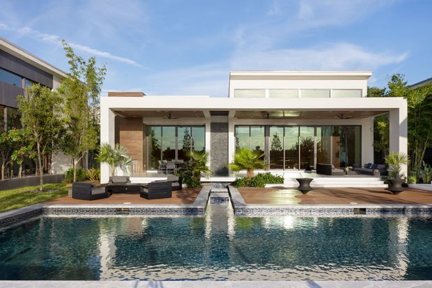 A contemporary modern residence viewed from the rear across a swimming pool. The house features a clean white rectilinear facade, broad covered outdoor terrace, extensive floor-to-ceiling glazing, dark stone-textured wall panels, timber decking, and a raised upper-level volume. The pool terrace includes outdoor lounge furniture and built-in planting, creating a polished indoor-outdoor entertaining area.