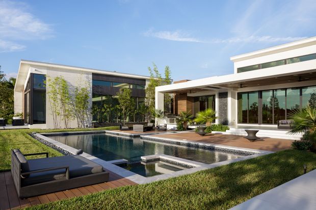 A bright contemporary multi-wing residence arranged around a landscaped courtyard and rectangular reflecting pool. The house features crisp white rendered walls, extensive dark-framed glazing, flat rooflines, timber accents, and a large covered outdoor entertaining terrace. The pool includes a shallow water feature and integrated stepping platform, with lawn, architectural planting, and outdoor lounge furniture surrounding it.