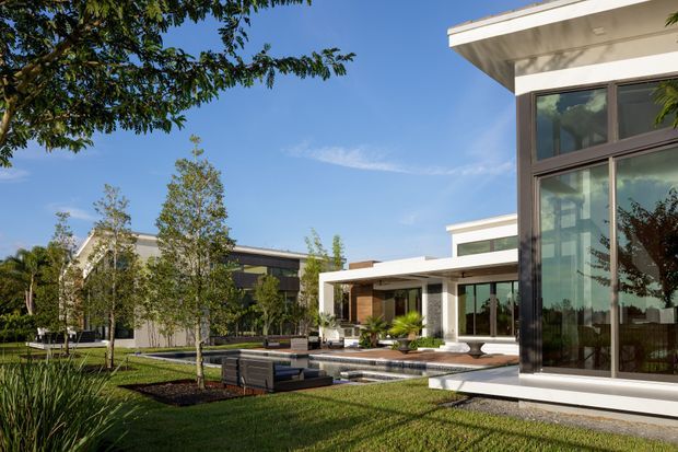 A spacious contemporary residential complex viewed from a landscaped garden, featuring crisp white flat rooflines, extensive floor-to-ceiling glazing, dark-framed windows, timber-toned wall panels, paved terraces, built-in outdoor seating, and a shallow linear water feature. Mature and newly planted greenery surrounds the modern low-rise structure under a clear blue sky.