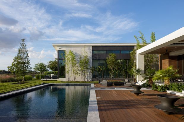Contemporary single-level residence with strong horizontal lines, pale rendered walls, extensive dark-framed glazing, and a deep covered outdoor entertaining area. A long rectangular reflecting or swimming pool borders a timber deck, with restrained landscaped planting and sculptural outdoor furniture creating a refined indoor-outdoor setting.