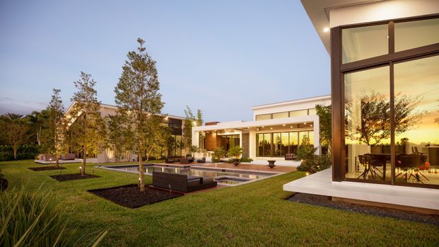 Wide exterior view of a modern single-storey residence at dusk, set within landscaped lawns and young trees. The home features strong horizontal volumes, flat roofs, expansive floor-to-ceiling glazing, warm interior lighting, and a generous paved terrace surrounding a long rectangular swimming pool. Outdoor lounge furniture and dining furniture are visible through the glazed living areas.