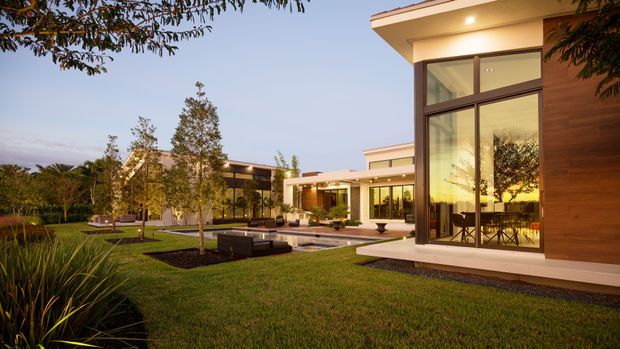 A dusk exterior view of a large contemporary residence arranged around a landscaped courtyard and shallow reflecting pool. The home features crisp flat rooflines, expansive floor-to-ceiling glazing, warm timber-clad walls, pale rendered surfaces, and a covered outdoor living area. Manicured lawn, planted garden beds, mature trees, and low dark outdoor seating surround the architecture.