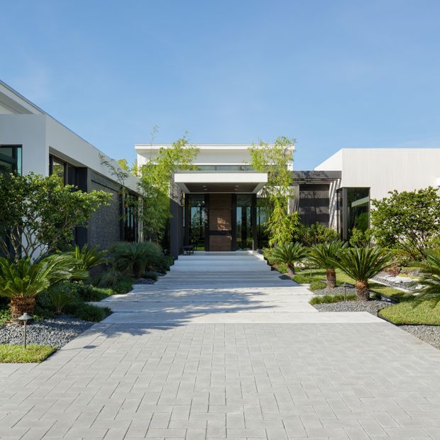 A contemporary single-storey residential entrance viewed from the front driveway. Broad pale grey pavers lead through a landscaped garden with palms, ornamental grasses and gravel beds to a recessed entry beneath a flat projecting canopy. The facade combines white rendered volumes, dark glazing and dark textured wall surfaces under a clear blue sky.