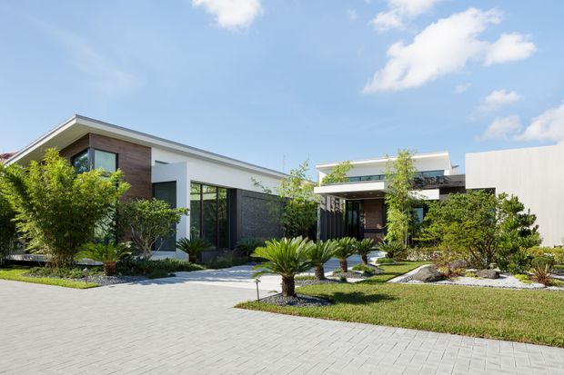 A contemporary single-storey residence with a low, flat roofline, crisp white and charcoal facade elements, extensive floor-to-ceiling glazing, and a covered entry. The home is set behind a broad light-grey paved driveway and carefully landscaped garden beds featuring sculptural palms, shrubs, rocks, and ornamental grasses. Bright blue-sky conditions and clean geometric forms give the exterior a polished, modern character.