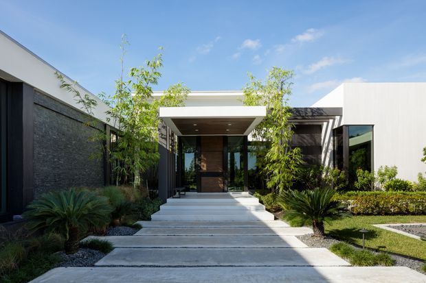 Modern single-storey residence viewed from the landscaped front approach. The facade combines clean white rendered volumes, dark vertical cladding, a textured dark stone feature wall, extensive glazing, and a broad flat-roofed entry canopy. Concrete stepping slabs lead through a structured garden with bamboo-like planting and cycads under a bright blue sky.