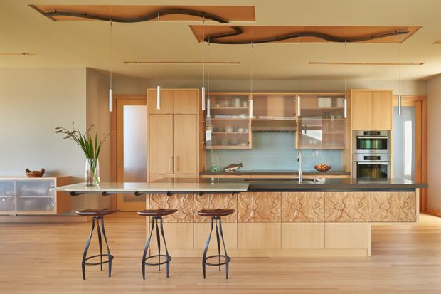 Open-plan contemporary kitchen with extensive light timber cabinetry, a long island bench with sculptural wood-grain drawer fronts, integrated appliances, glass-fronted upper cabinets, and three bar stools. The room has warm timber flooring, a dark stone-looking benchtop, pale walls, and distinctive sculptural timber ceiling features with suspended pendant lights.