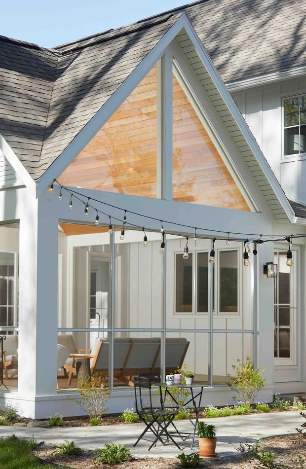 Exterior view of a bright, cottage-like home with a steep shingled gable roof, white trim and cladding, and a partially enclosed glazed porch or sunroom. The prominent front gable has warm timber shingle siding, while string lights, outdoor lounge chairs, a small café table, and planted garden beds create an inviting outdoor sitting area.