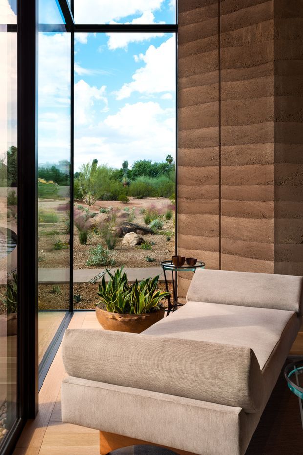 A refined seating nook inside a modern home, featuring a large upholstered chaise lounge beside full-height black-framed glazing. The room opens visually onto a landscaped, arid garden with native planting and rocks. A sculptural potted plant and a small round side table with cups add warmth, while a textured earthen-toned wall and timber flooring create a natural, calm atmosphere.