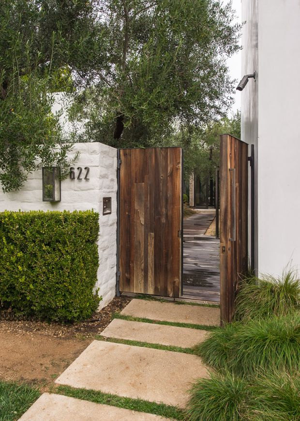A landscaped residential entryway viewed from outside, featuring a white textured masonry boundary wall marked with the number 622, tall vertical timber gates, a narrow timber-planked walkway beyond, clipped hedges, ornamental grasses, and mature leafy planting. The adjacent building has a plain white exterior wall with a small exterior light.