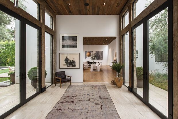 A refined residential entry hall framed by dramatic exposed timber posts and beams, with full-height black-framed glazing and glass doors opening to landscaped courtyards on both sides. The space has pale stone or porcelain flooring, a large muted patterned rug, white plaster walls, a richly stained timber ceiling, framed artwork, a sculptural chair, and potted greenery. The hall leads into a warm open-plan living or dining area beyond.