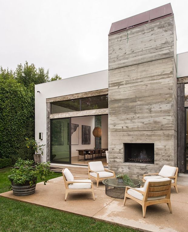 Modern courtyard exterior with a tall board-formed concrete fireplace chimney, white rendered walls, expansive dark-framed glazing, and a timber patio furnished with light wood and cream lounge chairs around a round outdoor coffee table. A leafy hedge and lawn frame the restrained contemporary composition.