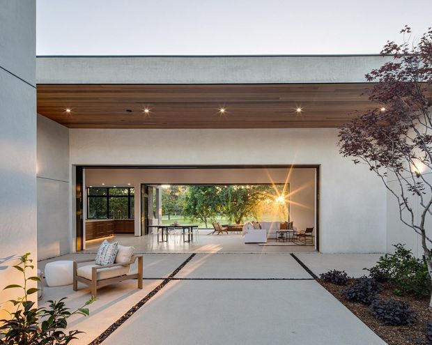 Contemporary courtyard-like outdoor living space framed by smooth pale rendered walls, a broad flat roof, and a warm timber-lined soffit with recessed lighting. Large open glazed doors connect the paved courtyard to a bright interior living and dining area, with minimalist seating, a dining table, landscaped planting, and a strong indoor-outdoor connection.