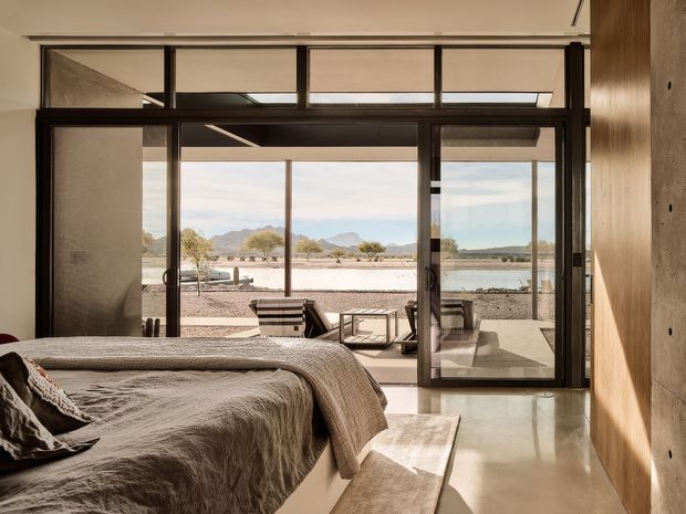 A serene modern bedroom with a low-profile bed in the foreground, opening through expansive dark-framed glazed doors to a covered terrace and an arid mountain landscape. The room features a polished pale floor, warm wood wall panelling, a neutral textured bedcover, and strong indoor-outdoor connection.