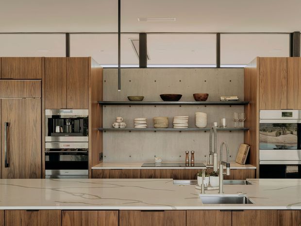 Contemporary kitchen featuring extensive warm timber cabinetry, a large pale stone island with integrated sink and tapware, and a concrete-look backsplash wall with open dark shelving displaying crockery and bowls. Built-in appliances are integrated into the cabinetry on both sides. A high clerestory-style glazed partition brings light into the space, while a slim dark pendant provides a vertical accent. The composition is clean, refined, and material-focused.