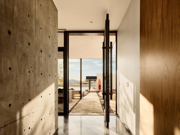 A sunlit modern interior entry or hallway opens through full-height black-framed glazing and a glass door onto a timber boardwalk and outdoor waterfront setting. The space features a raw exposed concrete wall with formwork marks, warm vertical timber wall panelling or joinery, polished pale flooring, crisp white walls, and strong indoor-outdoor visual connection. A distant body of water and mountains are visible beyond the deck.