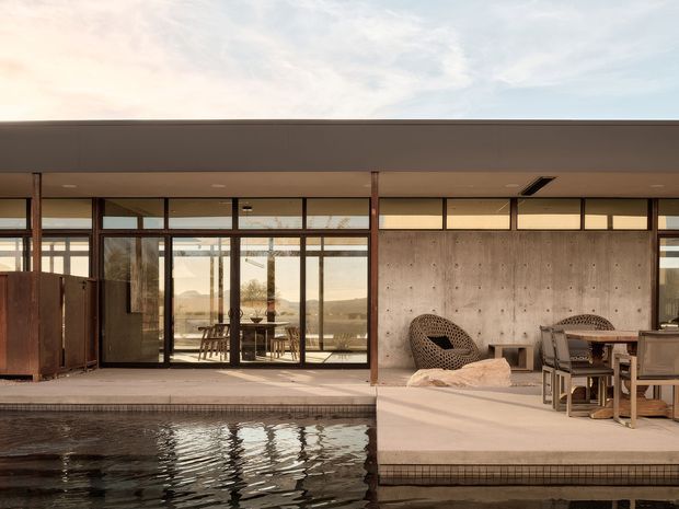 A refined modern indoor-outdoor entertaining space opening onto a swimming pool. The single-storey structure has broad floor-to-ceiling glazing, slim dark frames, timber posts, a deep flat roof overhang, and a raw concrete feature wall. A dining and lounge area sits on a pale tiled terrace beside the pool, with an expansive landscape visible through the glazing.