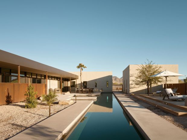 A low-slung modern residence arranged around a long, narrow reflecting pool or swimming pool. The scene features flat-roofed cubic volumes, extensive glazing, warm timber screening, concrete paving, gravel landscaping, desert-style planting, outdoor lounge furniture, and a clear blue sky with distant rocky terrain.