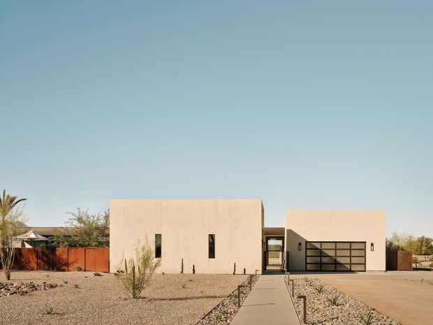 A low-profile modern residential property in a dry desert landscape. The composition shows two simple pale stucco volumes connected by a recessed glazed entry, with narrow vertical windows, a large dark-framed garage door, gravel landscaping, concrete paths, and warm timber fencing. The house has a restrained flat-roofed form and a muted neutral palette under a clear blue sky.