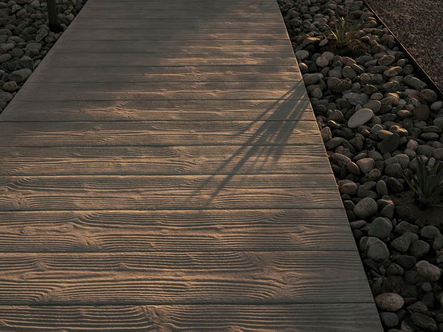 Close-up view of a narrow dark timber-look walkway or deck path bordered by rounded dark river stones and sparse drought-tolerant planting. The textured linear grain and warm low-angle light emphasize the surface finish, while a strong diagonal shadow crosses the path.