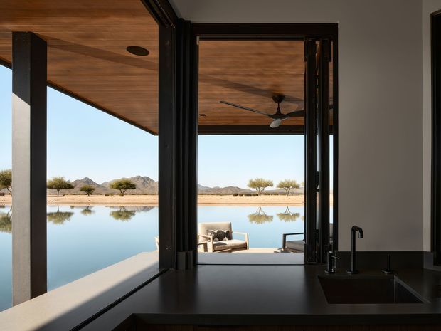 View from a contemporary kitchen through expansive black-framed glazing and open doors toward a covered outdoor lounge and reflective pool or pond, with a sparse desert landscape and mountains beyond. The kitchen foreground has a dark benchtop, integrated sink and black tap, while the soffit and ceiling are clad in warm timber boards. Strong indoor–outdoor connection and long water views dominate the composition.