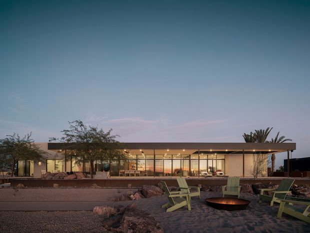 Twilight exterior view of a low, single-storey modern residence with a broad flat roof and extensive floor-to-ceiling glazing. The house opens onto a landscaped rocky yard with gravel, desert-style planting, lounge chairs and a circular fire pit, creating an indoor-outdoor entertaining setting.