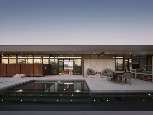 Wide dusk view of a low, flat-roofed contemporary residence arranged around a dark reflecting swimming pool. Full-height glazed doors open onto a pale paved terrace, with timber privacy screens, outdoor dining furniture, lounge chairs, and a built-in outdoor fireplace or wood-storage feature visible along the facade.