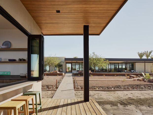 A contemporary single-storey courtyard-style residence viewed from a covered timber deck. The foreground shows an outdoor kitchen or servery with stools, timber decking, a slatted timber ceiling, dark structural posts, and open glazing. A straight paved path leads through a dry, landscaped courtyard toward a low, glass-fronted building with broad roof overhangs. The architecture has a restrained desert-modern character with strong indoor-outdoor connections and warm natural materials.