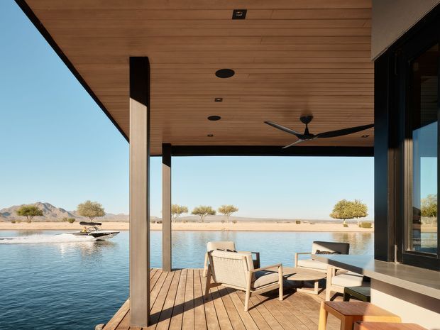 A covered outdoor terrace or deck overlooking a swimming pool, with timber decking, a timber-lined ceiling, slender structural posts, black-framed glazing, an outdoor ceiling fan, and contemporary lounge furniture. The pool and arid landscaped setting extend toward low mountains in the background.