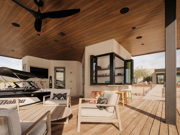 A covered outdoor living area with a warm timber-lined ceiling, recessed lights, and a large black ceiling fan. The space has timber decking, a built-in bar or serving window with black-framed bi-fold glazing, light upholstered lounge furniture, and a dining table. An adjacent open area and boat are visible beyond the covered zone.