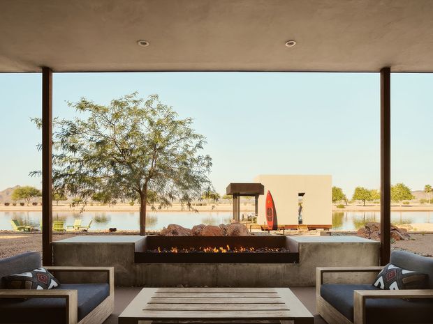 A covered outdoor lounge overlooking a landscaped water feature, with two upholstered seating areas facing a long built-in linear fire pit. The space has a broad concrete ceiling with recessed downlights, slim dark structural posts, concrete fire-pit surround, and a low timber slat coffee table. The view includes rocks, trees, water, and a minimalist pavilion-like structure beyond.