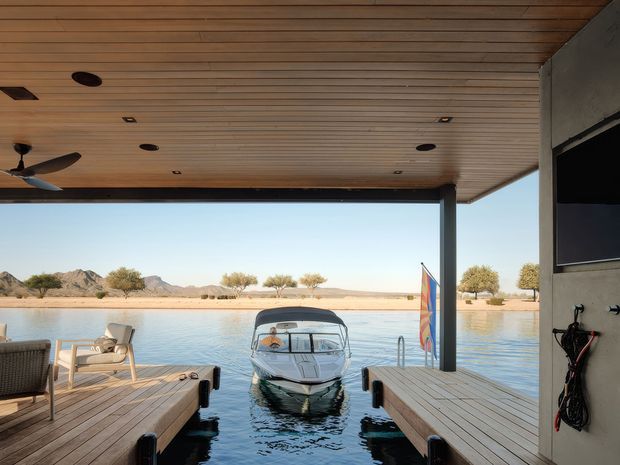 Covered waterfront boat dock and outdoor entertaining area with timber decking, a warm timber-lined ceiling, recessed lighting, ceiling fans, and a central motorboat moored between two floating platforms. A calm lake, arid mountains, and sparse shoreline vegetation are visible beyond.