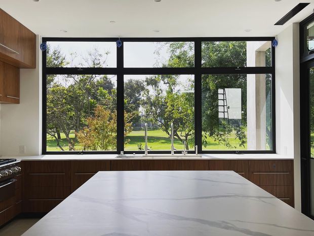 Bright contemporary kitchen with dark timber cabinetry, a large pale stone island bench, stainless steel sink and tapware, and expansive black-framed windows overlooking a leafy garden. The white ceiling includes recessed downlights and a linear ventilation or lighting slot.