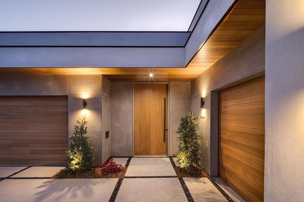 A contemporary residential entry courtyard viewed from outside, featuring a warm timber front door, matching timber garage doors, smooth grey rendered walls, a timber-lined soffit, integrated wall lighting, and large pale concrete pavers separated by dark gravel joints. Slim planted beds frame the entry, creating a refined modern arrival space.