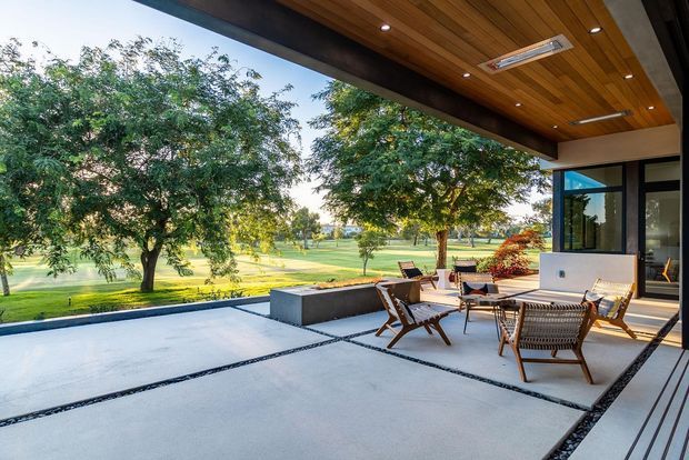 A covered outdoor terrace overlooking a manicured green landscape. The expansive pale concrete patio is divided by dark linear gravel or stone joints, with timber-framed outdoor seating and a low built-in planter or bench. A broad timber-lined ceiling with recessed downlights and a skylight-like roof window shelters the space, while large black-framed glazed doors connect it to the contemporary interior.